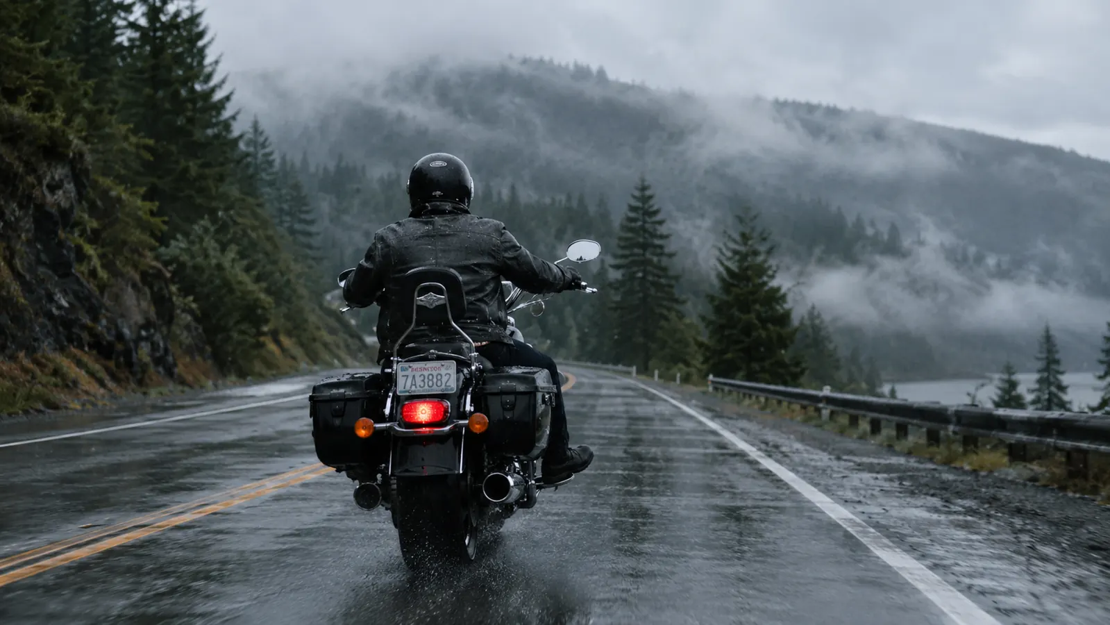 A motorcyclist riding along a wet rural road with a misty treeline in the rain, seen from behind.
