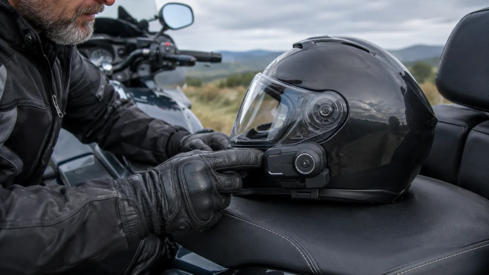 A rider's gloved hand reaching toward a small intercom unit mounted on the side of a motorcycle helmet