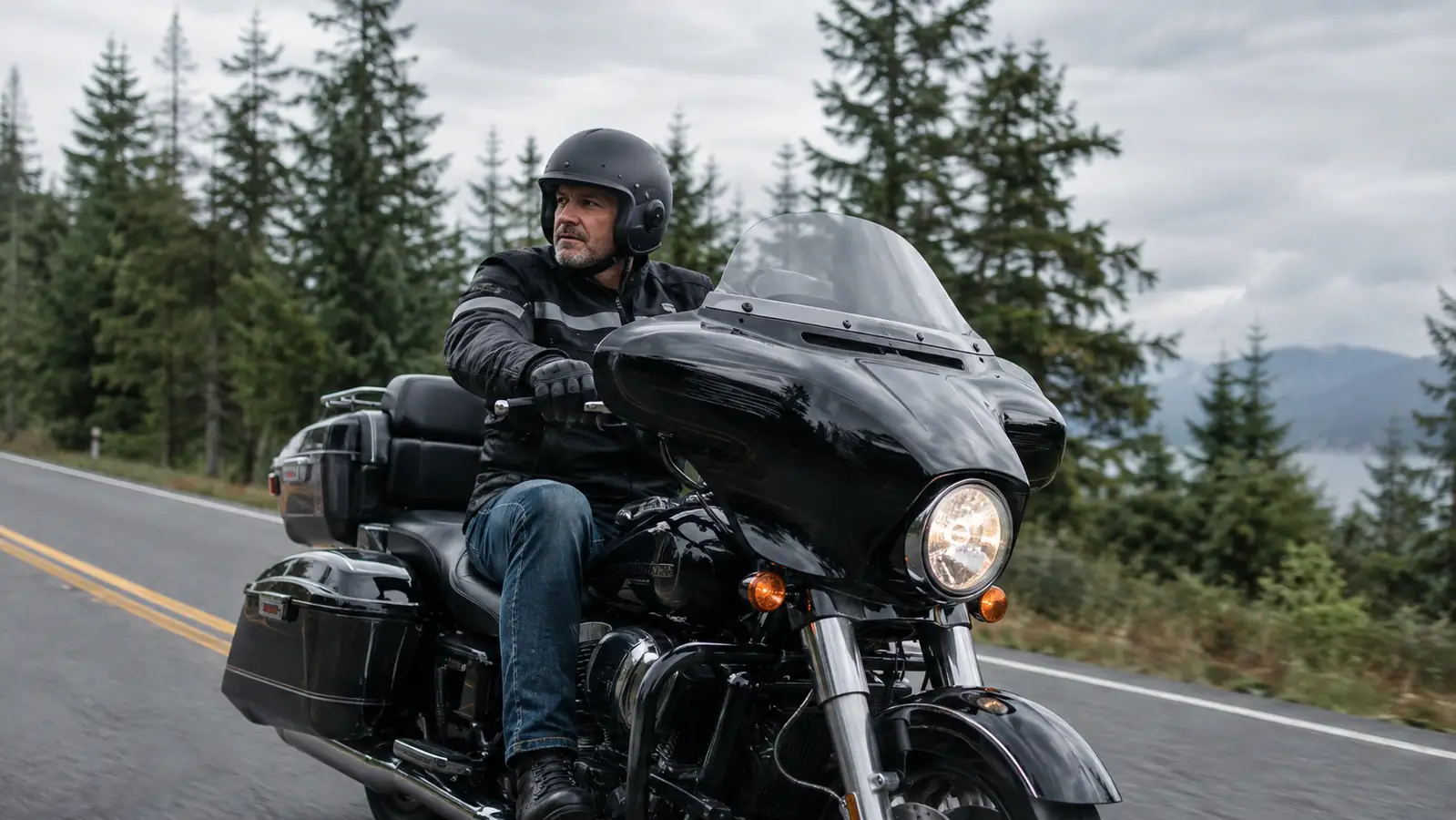A rider in a half helmet and riding jacket seated on a large touring motorcycle on a two-lane road, evergreen trees lining the road