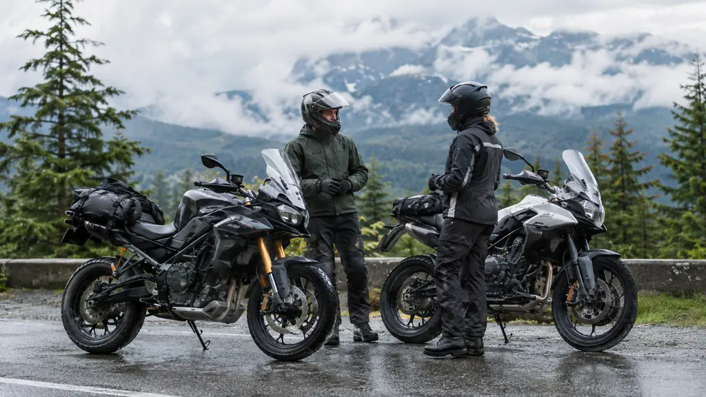 Two motorcycles parked side by side at a rest stop, two riders in helmets standing and talking beside their bikes, rain gear visible, evergreen trees and low clouds behind